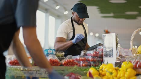 un hombre negro con una camiseta blanca y un delantal negro en una gorra negra clasifica las verduras en el mostrador en una tienda de comestibles moderna. los empleados de una gran tienda moderna colocan las mercancías en el counter durante su servicio