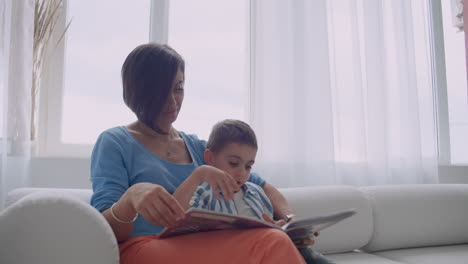 madre amorosa enseñando a su hijo a leer sosteniendo un libro hablando en el sofá en casa