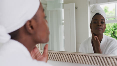 Mujer-Afroamericana-En-Bata-De-Baño-Aplicando-Crema-Facial-Mientras-Se-Mira-En-El-Espejo-En-Casa