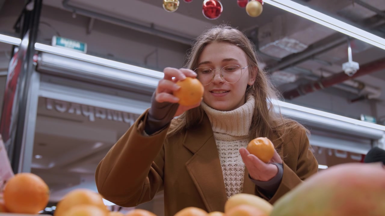 Donna bianca che ispeziona mandarini da vicino. Inquadratura ravvicinata lifestyle con dolcevita e occhiali, espressione calda, mani che tengono due mandarini, focus su buccia e consistenza, illuminazione soffusa del mercato.
