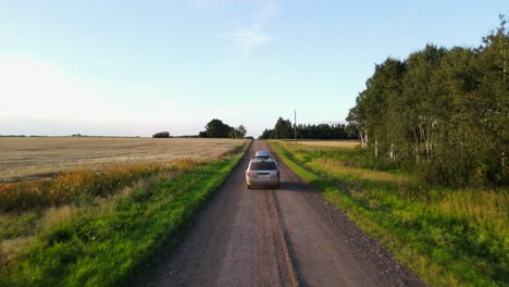 aerial tracking shot of a silver minivan driving along a dusty dirt road during sunset in the canadian countryside