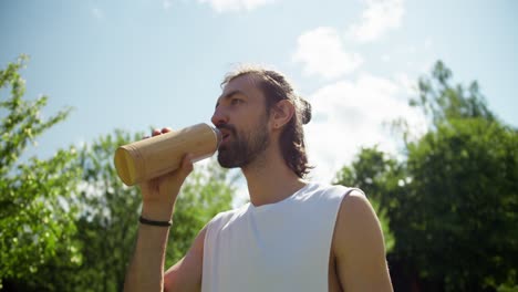 An-overview-of-a-brunette-guy-in-a-white-T-shirt-who-drinks-water-in-nature-against-the-background-of-green-trees-and-a-blue-sky.-Rest-and-break-after-playing-outdoor-sports