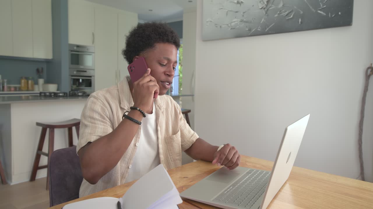 Adult male taking work call, holding magenta phone, typing on laptop at dining table with papers