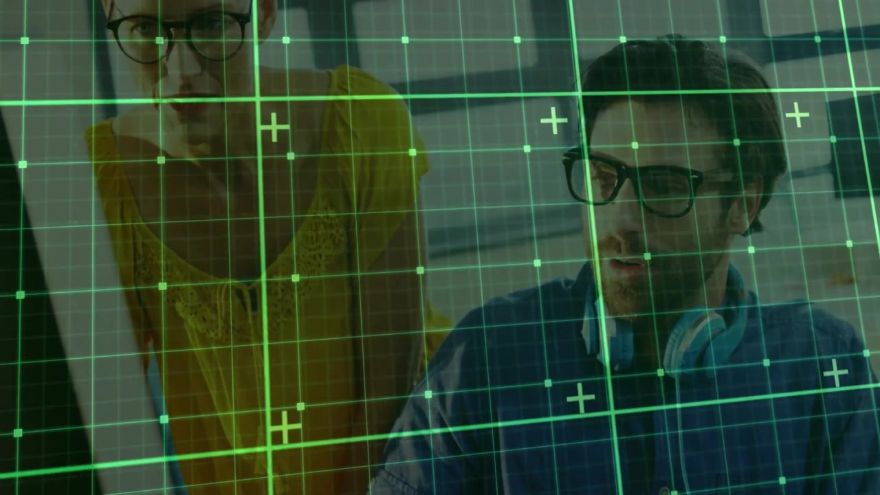 Coworkers leaning over desk and tapping tablet, reviewing tech design with green grid overlay
