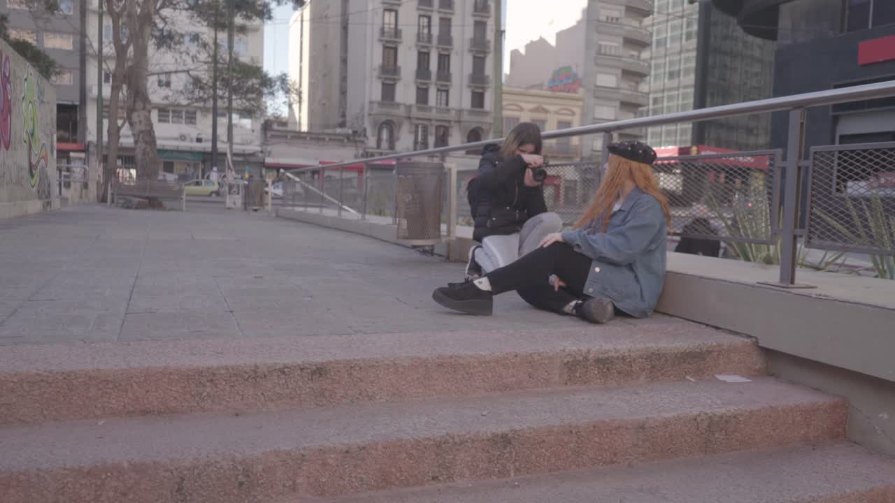 Two friends have fun taking pictures in the street.  Redhead woman model poses sitting on the sidewalk and she smiles at her friend's directions