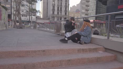 two friends have fun taking pictures in the street.  redhead woman model poses sitting on the sidewalk and she smiles at her friend's directions