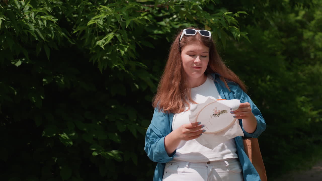 Young girl in casual outfit walking through lush green alley holding embroidered fabric, smiling proudly at her creative work while dancing lightly in sunlight