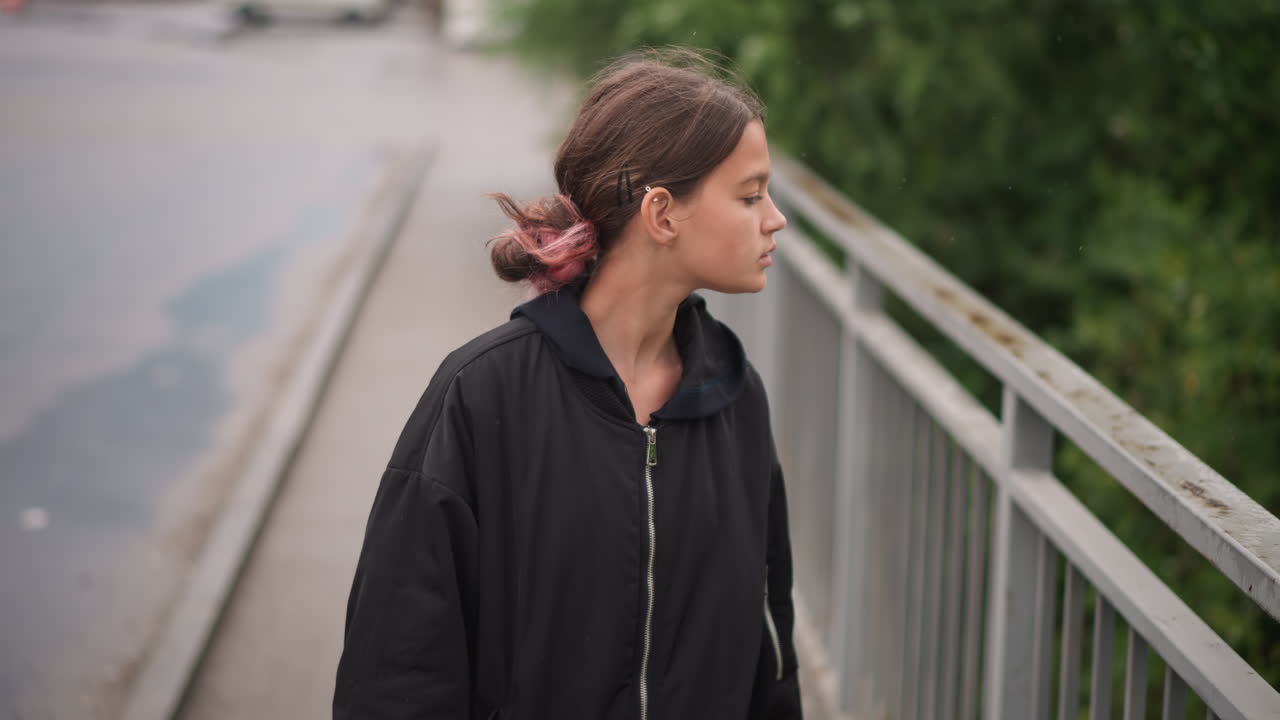 Giovane donna su un ponte sotto la pioggia, che indossa una felpa con cappuccio nera, in piedi vicino a una ringhiera bagnata e pozzanghere, sguardo contemplativo verso lo skyline, cielo coperto e fogliame verde, pioggerella leggera lascia goccioline nei capelli.