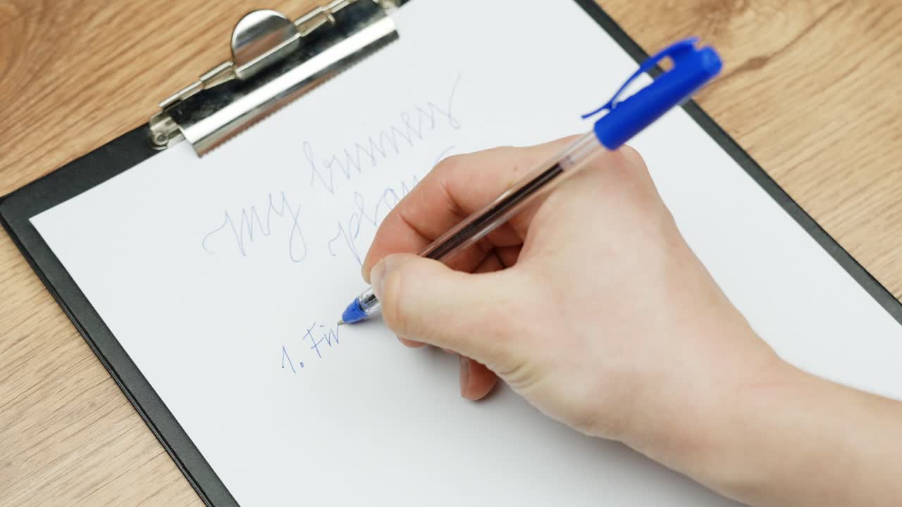 Close-up of a hand holding a blue pen writing a numbered list on a clipboard with "my business goals" at the top. Captures a moment of planning, motivation, and seasonal aspiration.