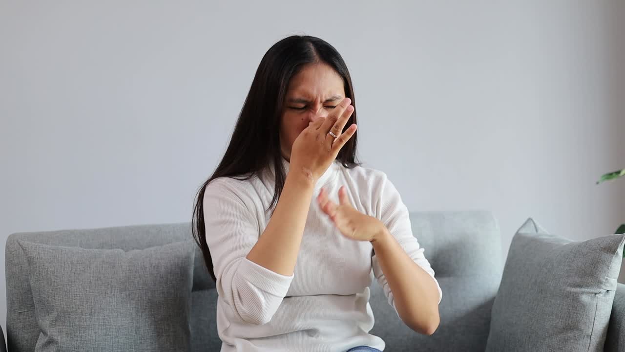 Giovane donna asiatica seduta sul divano di casa, che sente un odore nauseabondo e disgustoso, un lezzo intollerabile, trattenendo il respiro con le dita sul naso. Cattivo odore.