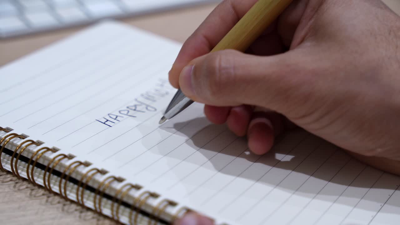 Close-up of a hand writing an event checklist in a spiral notebook on a wooden desk, with keyboard in soft indoor light and shallow depth of field for planning and organization concepts