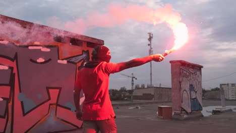 young man in balaclava with red burning signal flare on the roof with graffiti background, slow motion