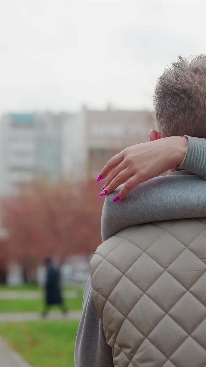 Un soggetto maschile rimane fermo in un parco urbano. Gli individui mantengono le posizioni sotto un cielo urbano nuvoloso. Lo sfondo mostra una persona sfocata che si allontana con vestiti e scarpe scuri.
