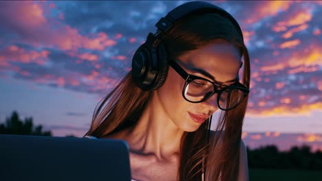 mujer joven enfocada con auriculares trabaja en su portátil al aire libre, con vibrantes colores del atardecer reflejados en sus gafas, creando una atmósfera cautivadora y serena