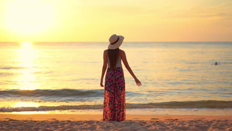 Back-of-Stylish-Petite-Female-Standing-Alone-on-Sandy-Beach-on-Golden-Hour-and-Watching-Sunset-Over-Tropical-Sea-Horizon