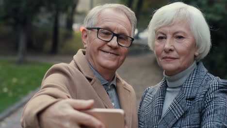 Front-View-Of-Elderly-Couple-Posing-And-Smiling-To-The-Camera-While-Taking-Selfie-Photo-With-Smartphone-In-The-Park-At-Sunset