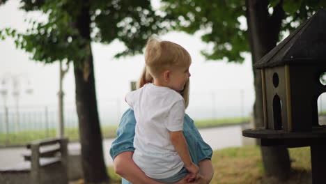 a mother lovingly carries her child close to a birdhouse, allowing the child to look closely at it, with trees and bench behind them