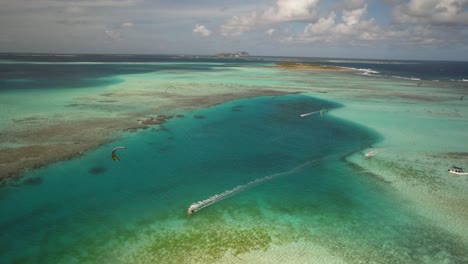 Kitesurfers-gliding-over-a-clear-turquoise-sea-with-a-coral-reef,-aerial-view