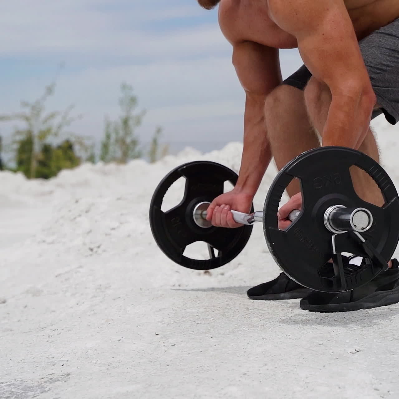 Athlete is taking heavy barbell on the natural background. Sporty man in shorts and trainers starting his workout to lift barbell outdoors. Slow motion.