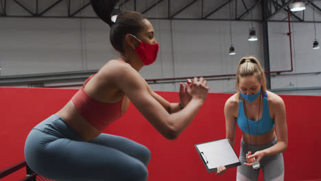 Mujer-De-Raza-Mixta-Saltando-Sobre-Una-Caja-Usando-Máscara-Facial-En-El-Gimnasio,-Sincronización-De-Mujer-Caucásica