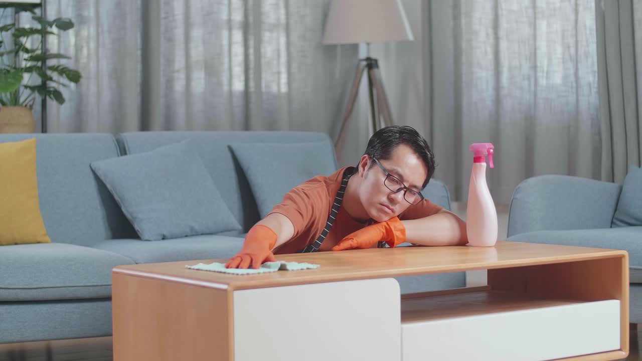 Bored Asian Male Housekeeper With An Apron Cleaning The Table By The Spray At Home