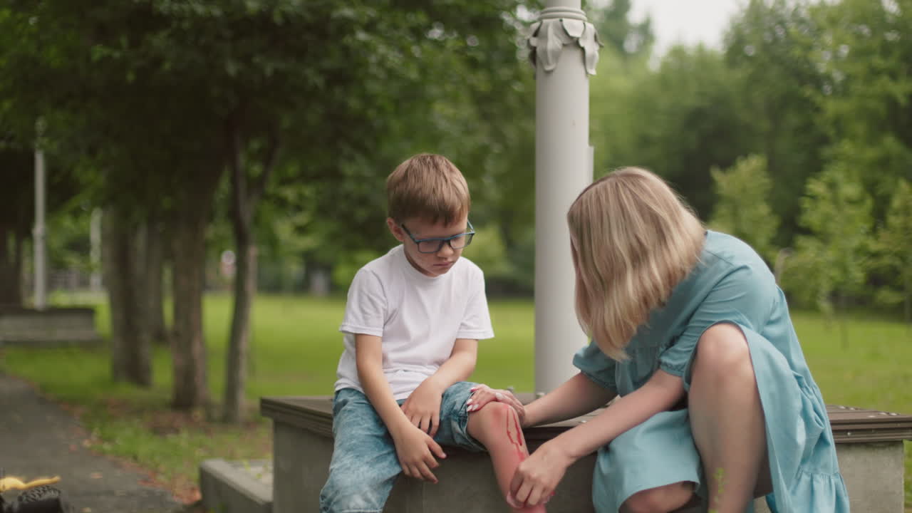 una madre se arrodilla al lado de su hijo en una tranquila carretera del parque, limpiando suavemente su herida en la pierna mientras él mira hacia abajo, visiblemente triste