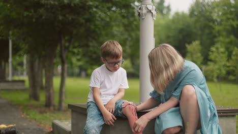 a mother kneels beside her son on a quiet park road, gently cleaning his leg wound while he looks down, visibly sad