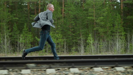 a man wearing a grey suit jacket and blue jeans is captured mid-stride as he runs along railway tracks in a forested area, the lush greenery and electric poles in the background