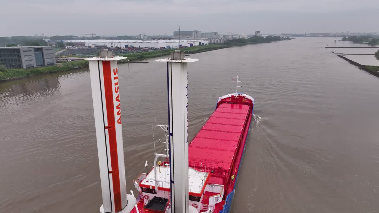 A cargo ship equipped with rotor sails navigating a river past an industrial area.