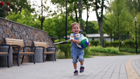 happy caucasian toddler runs by the road holding a ball. beautiful smiling baby runs up to camera. low angle view.