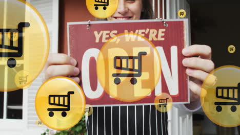 animation of shopping cart icons over smiling caucasian woman holding yes we are open text board