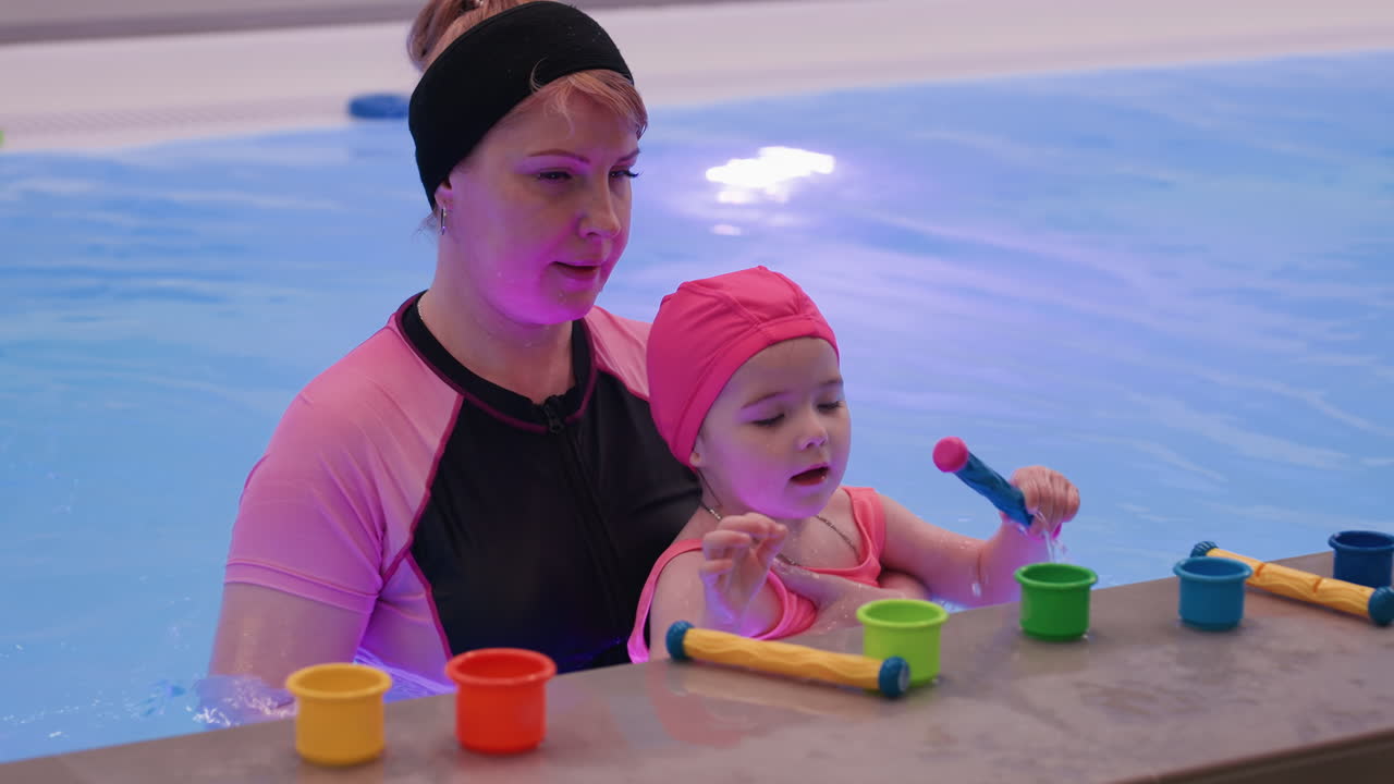 어린 소녀가 수영장 바닥에 멋진 드럼 스틱을 놓고 있어요. 조심스러운 손길로 장난감을 물가 근처에 놓고, 햇빛이 비치는 물보라가 반사되는 모습이에요. 손과 눈의 협응력에 초점을 맞추고, 차분한 분위기의 어린 시절 수영 레슨 느낌을 담았어요.