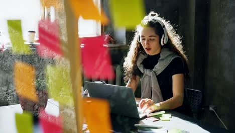 creative young businesswoman is listening to music in headphones dancing and singing while working at desk with laptop in modern office. glass with colored stickers in foreground.