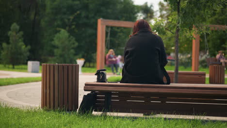 vista trasera de una dama sentada en un banco del parque, vestida con abrigo y botas negras, rodeada de árboles verdes exuberantes, atmósfera al aire libre relajada en un parque urbano, vista borrosa de dos personas en sillas giratorias