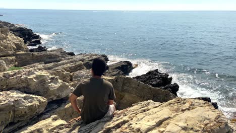 young man sitting on the rocks enjoying the view of ocean at summer in saint-jean-cap-ferrat, france