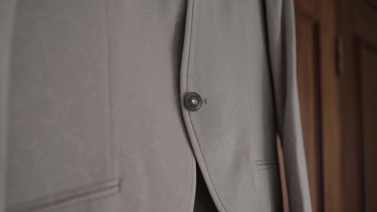 Primo piano del bottone e della tasca frontale di una giacca da completo beige su tessuto sartoriale.