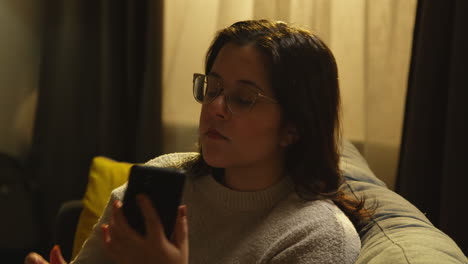 mujer joven en casa sentada en el sofá con teléfono móvil desplazándose por internet o redes sociales por la noche 1