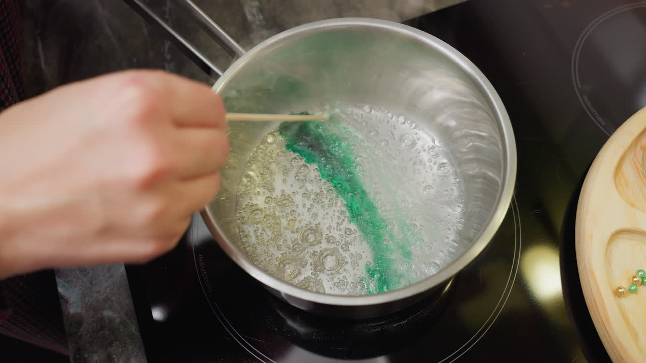 Close-up of person stirring in pot with green colorant, steam rising from liquid, beads of different colors green and golden on wooden tray in background, cooking or food preparation process