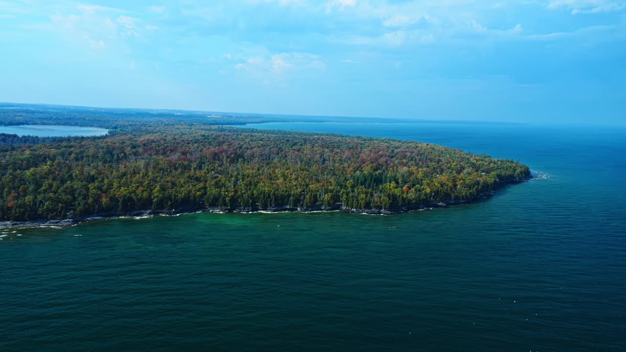 The forested tip of Door County reaches into Lake Michigan, its trees glowing with early autumn hues above turquoise waves and rocky shores under a clear blue sky
