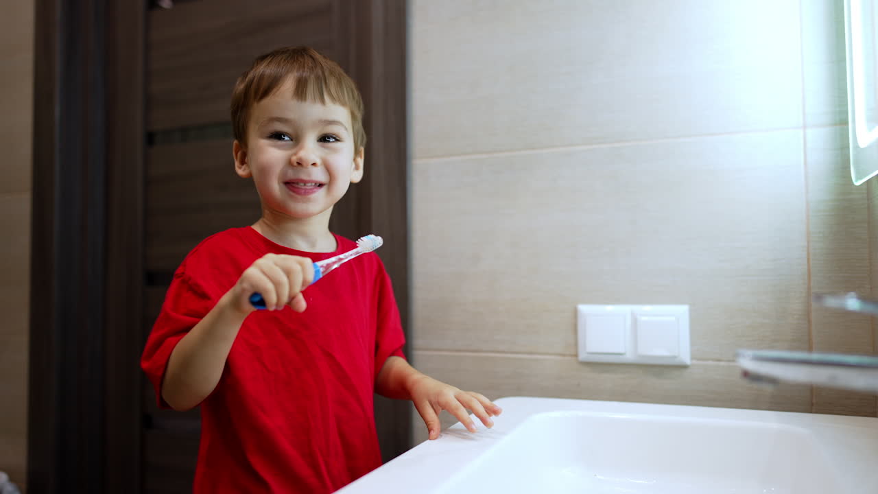 Lovely baby in red t-shirt holds a toothbrush. Kid switches on electric toothbrush and starts cleaning teeth.