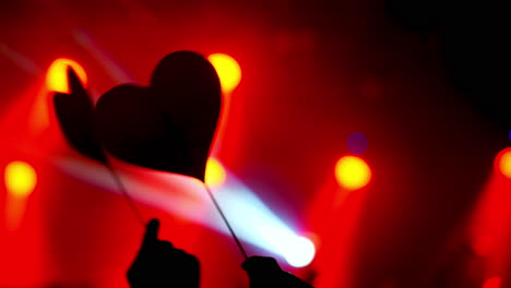 silhouetted hands holding heart shaped paper cutouts sway rhythmically in a dimly lit concert venue, illuminated by vibrant red and blue spotlights