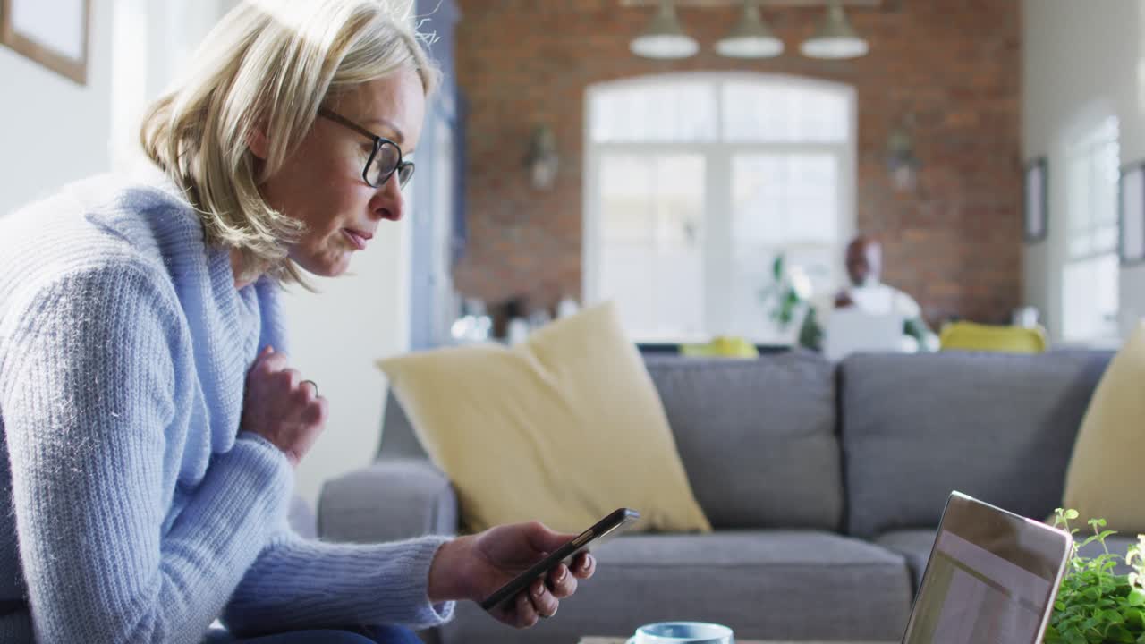 Worried senior caucasian woman in living room sitting on sofa, using smartphone and laptop