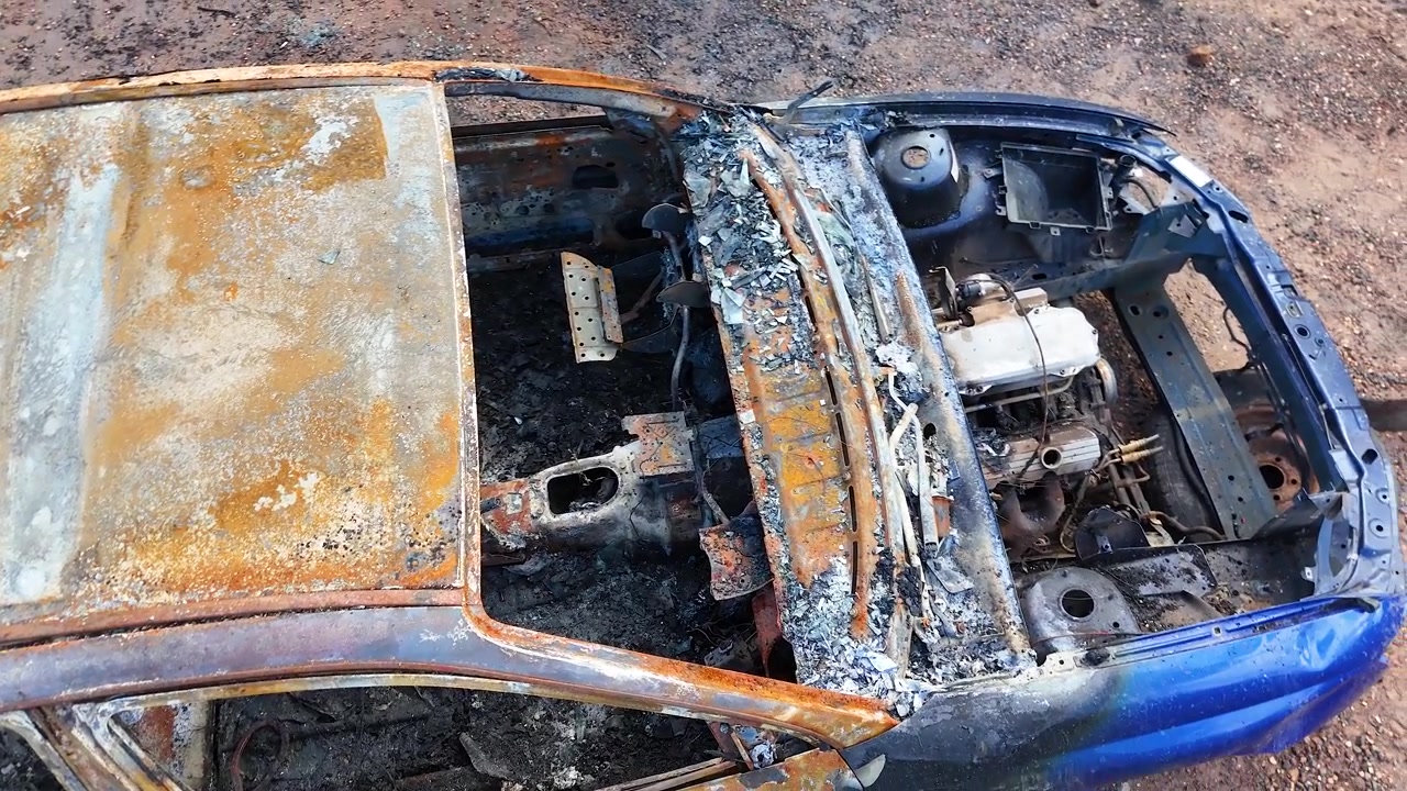 Top-down drone view tracking over a rusted, fire-gutted car wreck in Goonoo Forest, Australia.