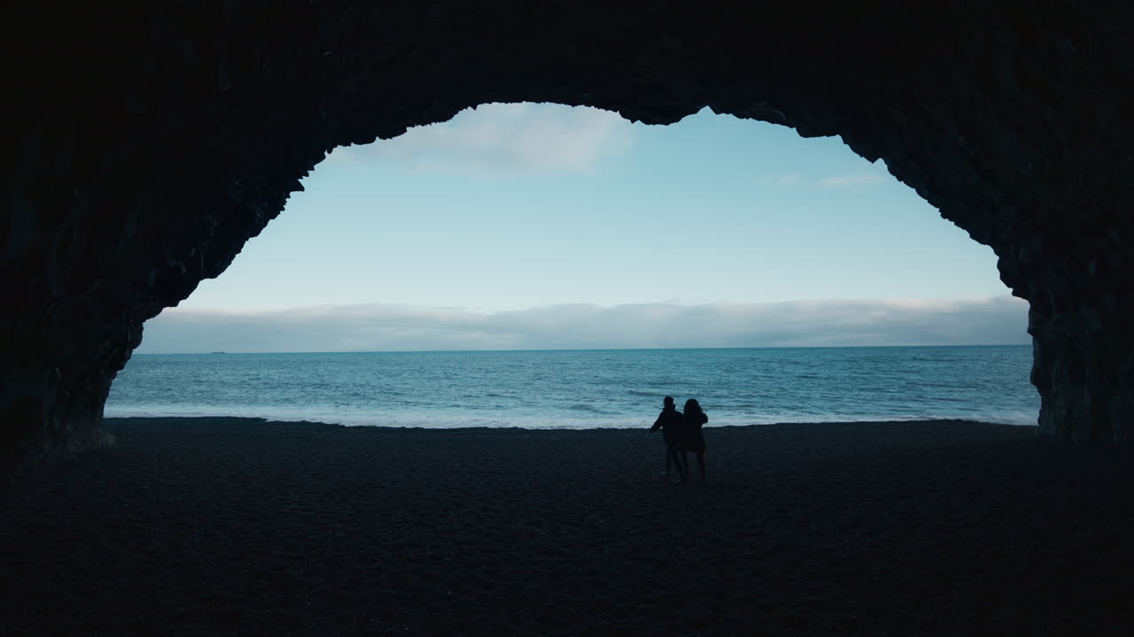 View of Reynisfjara Beach, from a cave, couple walking in distance, moody silhouette, high contrast, wide angle, Iceland 02