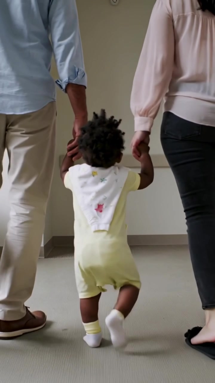 Video verticale: Genitori che iniziano a far camminare il bambino piccolo in tutina con bavaglino che fa i primi passi lungo il corridoio verso la porta