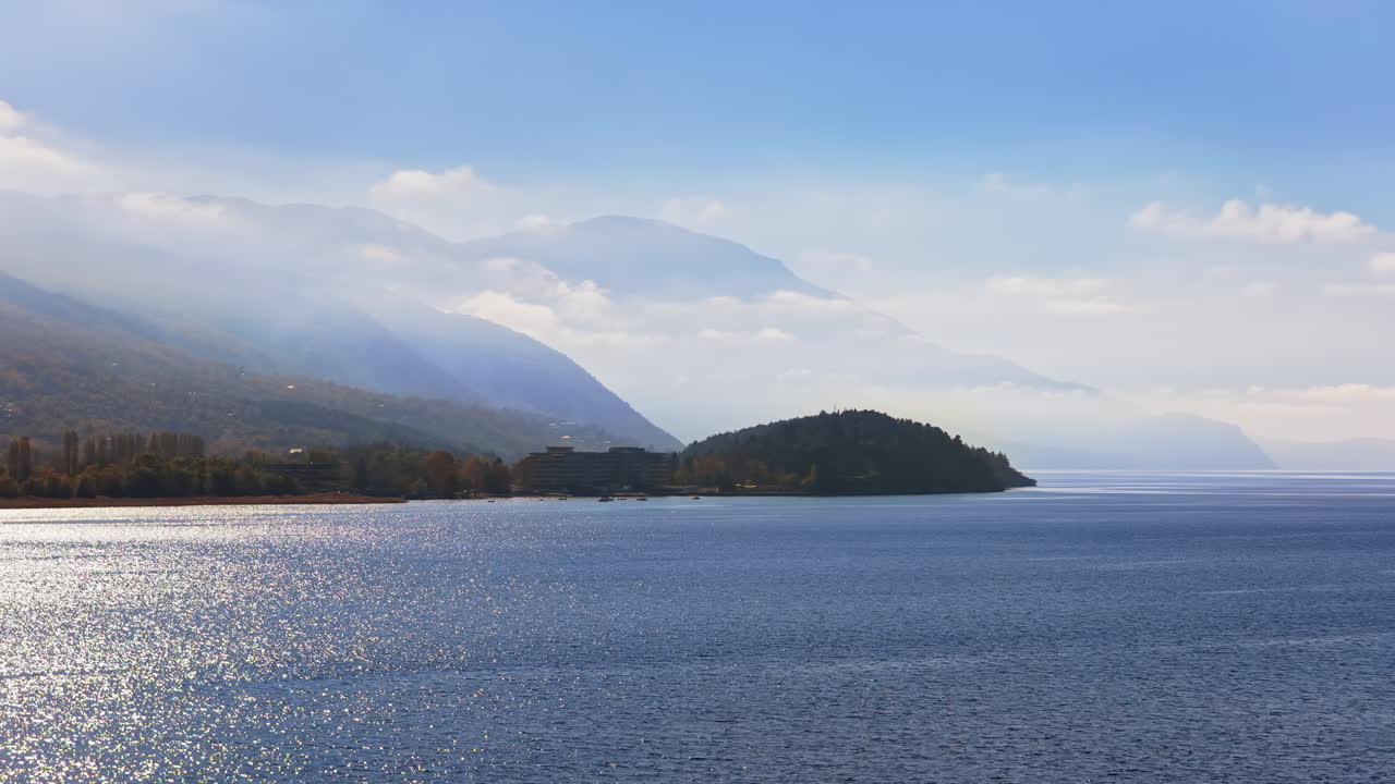 Aerial drone view of the surface of Lake Ohrid, with the mountains gently covered in morning mist and a distant peninsula completing the peaceful landscape