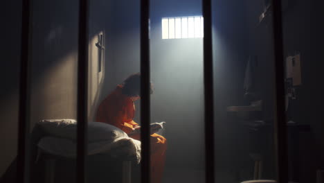inmate reading holy bible on bed in prison room with barred window