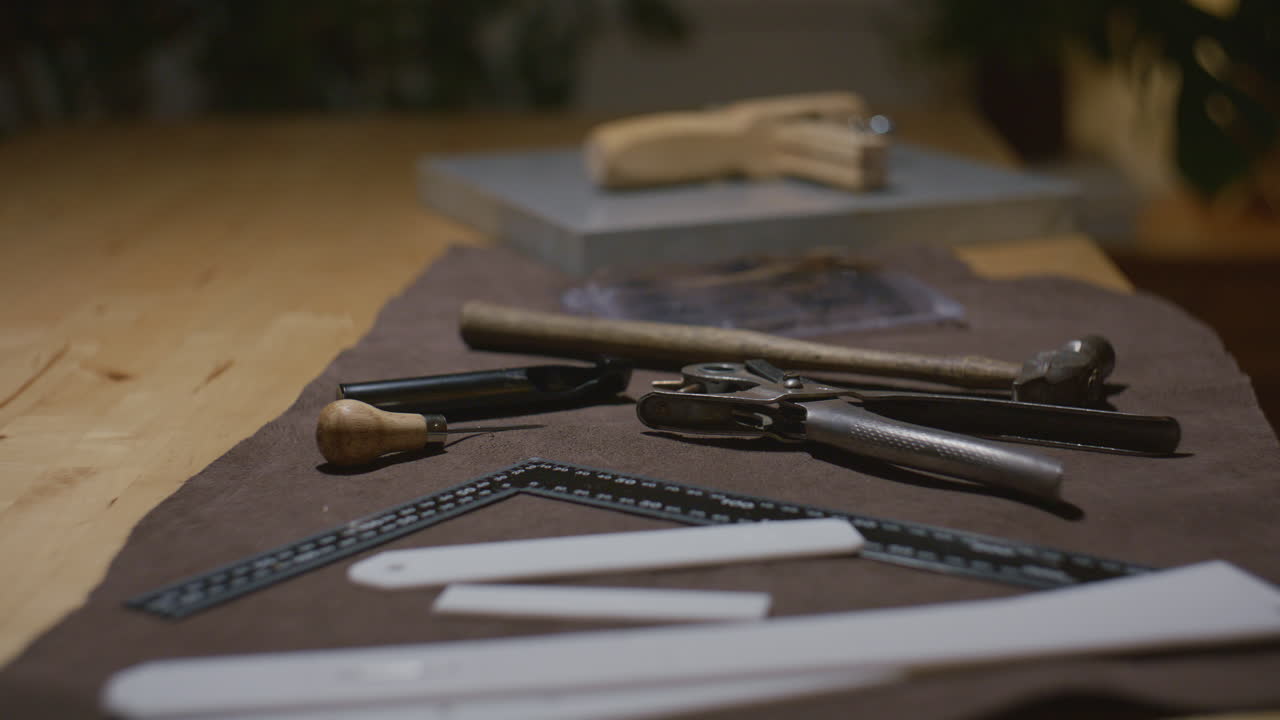Various leather crafting tools, including a hammer, ruler, and cutting instruments, placed on a brown leather surface in a workshop.