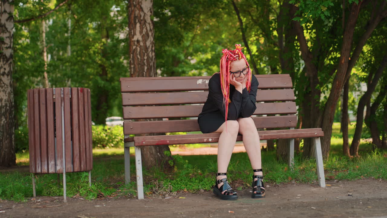 Сидящая женщина с неоновыми дредами, обхватившая голову руками за щеки, залитый дневным светом парк, деревянная скамейка, мусорный бак поблизости, задумчивое выражение лица, медленное дыхание, созерцательная пауза