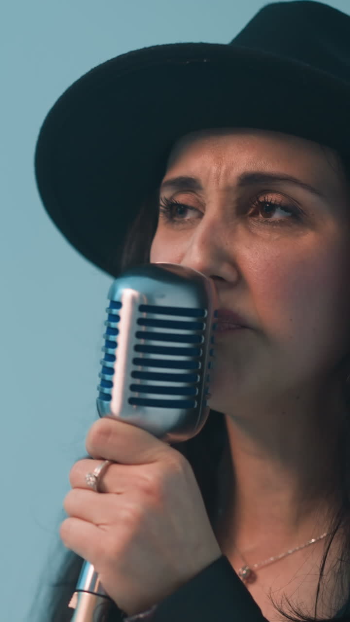 검은 모자와 가죽 재을 입은 열정적인 가수가 파란색 배경에 비추어 빈티지 마이크로 노래하는 클로즈업 뷰.